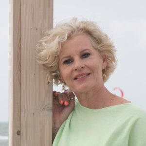 Karin de Jong, HSP-coach, leunend tegen een houten paal bij het strand, met een ontspannen en toegankelijke uitstraling.